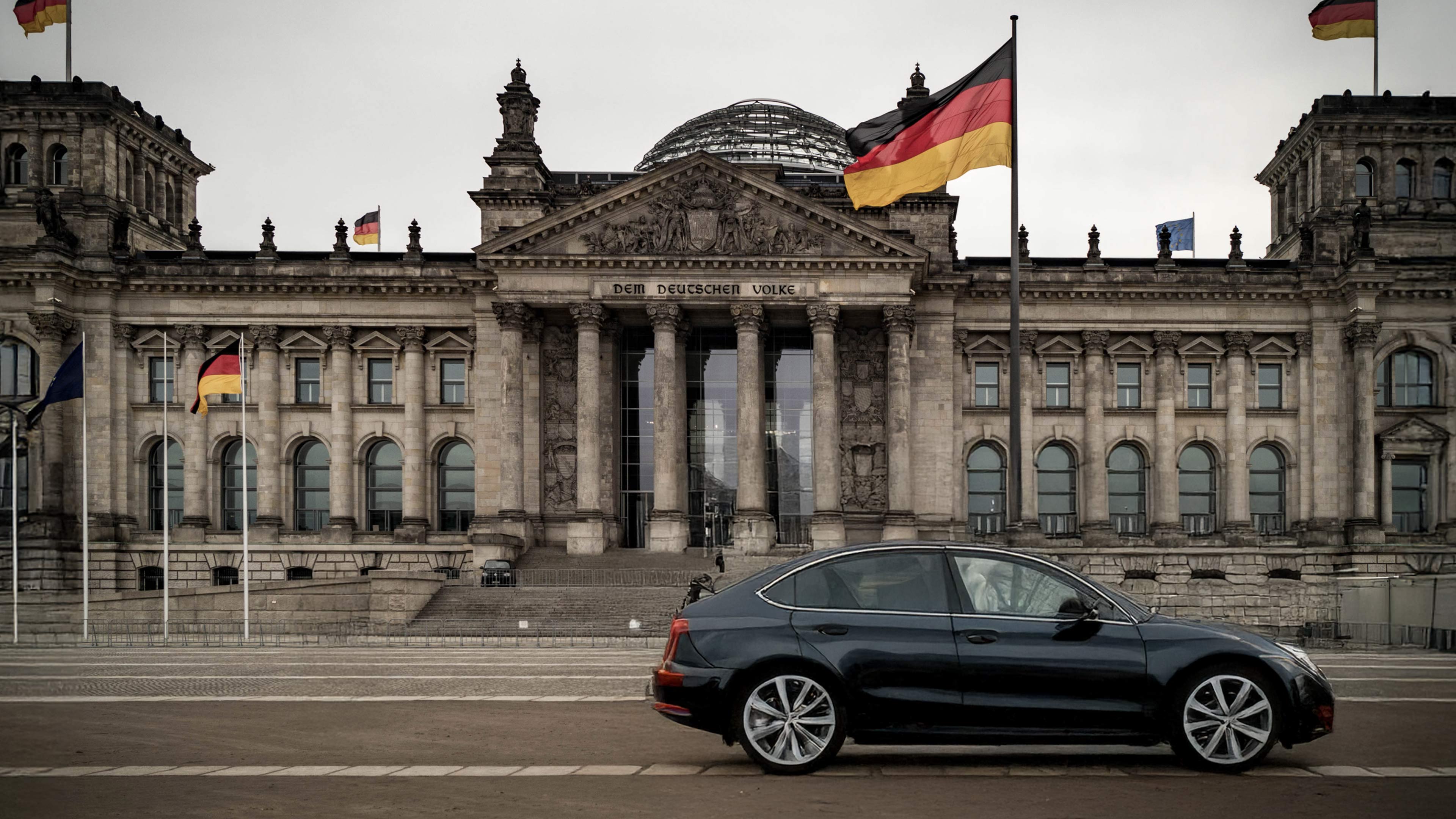 Schwarzes Auto vor dem Reichstagsgebäude in Berlin, mehrere deutsche Flaggen sichtbar. Symbolisiert: Produkt ist in Deutschland zugelassen.