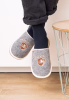 Prachtkerl Filzpantoffeln hergestellt aus echtem italienischen Wollfilz baumelnd ein Paar graue Filzpantoffel mit der hellbraun-weißen Stickerei eines Löwenkopfes in einem Wappen mit dem Schriftzug Prachtkerl darüber. Außen hell eingefasst