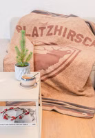 liegend auf einem Sofa eine hellbraune Kuscheldecke mit braunem Wappen und röhrendem Hirsch sowie dem Schriftzug Platzhirsch oben drüber. Oben und unten dunkelbraune und graue Streifen als Abschluss der kurzen Seiten