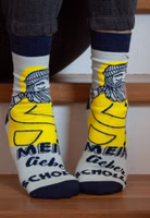 jemand sitzend auf Treppenstufen mit einem Paar hellgraue Mein lieber Scholli Socken mit Fischer Motiv in leuchtend gelbem Friesennerz sowie dem dunkelblauen Schriftzug Mein lieber Scholli. Dunkelblau abgesetzter Bündchen-, Fersen- und Zehenbereich