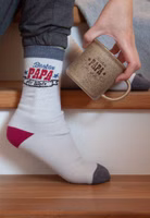 jemand sitzend auf Treppenstufen mit einem Paar hellgrauen Sport-Socken mit dunkelgrau abgesetztem Bündchen sowie Zehenbereich und dunkelrot abgesetztem Fersenbereich. Dazu im oberen Knöchelbereich in blau und rot der Bester Papa der Welt Schriftzug