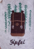 auf hellbeigem Filz eine Stickerei eines Wanderrucksacks in Brauntönen und grünen Tannenbäumen sowie dem Schriftzug Gipfel
