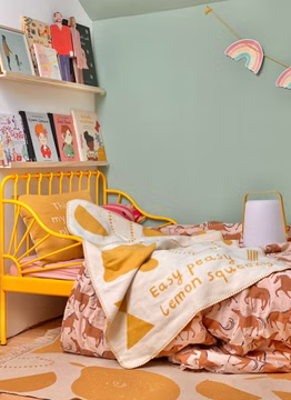 eine Baby-Kuscheldecke in beige und senf mit dem Motiv "Easy Peasy" liegend auf einem gelben Kinderbett im Kinderzimmer