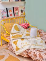 eine Baby-Kuscheldecke in beige und senf mit dem Motiv "Easy Peasy" liegend auf einem gelben Kinderbett im Kinderzimmer