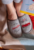 auf einer Decke ein Paar graue Filzpantoffeln rechts mit dem Schriftzug in dunkelgrau und rot "Let the adventure begin" und links die Stickerei eines Fahrrades und Bullies in rot weiß.