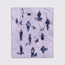 eine helllila-farbene Kuscheldecke mit dem dunkelblauen Motiv von verschiedenen Hundebesitzern mit deren Hunden zum Teil an der Leine liegend auf einem Sessel
