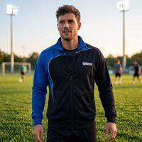 A man is standing on a soccer field, wearing an OMKA tracksuit jacket. A soccer goal and players are in the background.